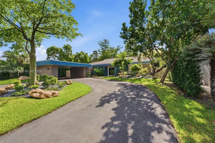 Paved circular driveway leading to a covered carport, mature trees, manicured lawn, and established landscaping