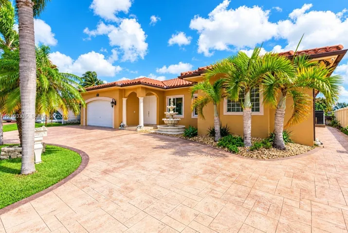 Stucco exterior featuring a red tile roof, multi-car garage, decorative stone fountain, and patterned concrete driveway