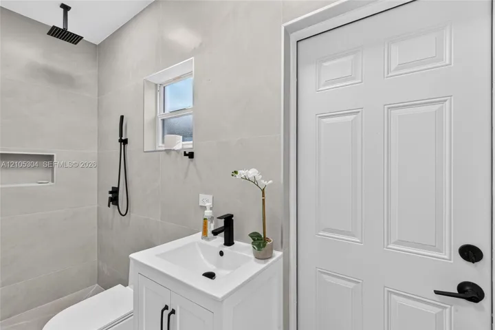 Full-height neutral wall tiling, walk-in shower with rainfall showerhead and built-in wall niche, white vanity with rectangular basin and matte black fixtures, and six-panel interior door with matte black hardware