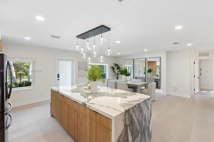 Kitchen island with stone waterfall countertop and natural wood-finish cabinetry, illuminated by a multi-pendant light fixture, complemented by light-toned wood-finish flooring and recessed ceiling lighting
