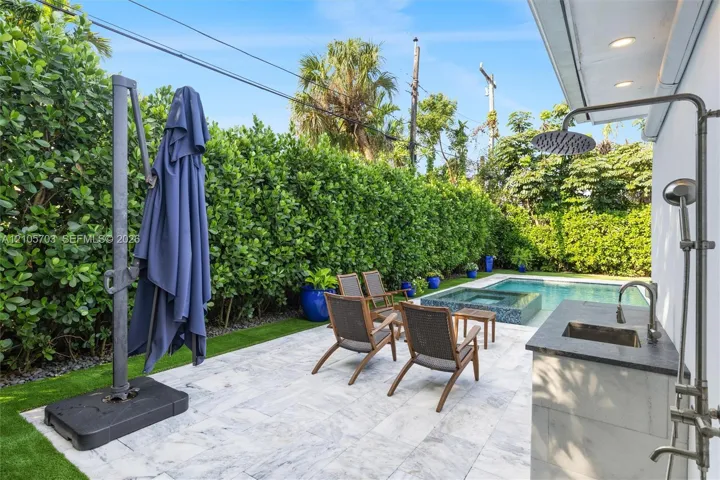 Outdoor patio featuring natural stone paving, an integrated wet bar with a dark stone countertop, a wall-mounted shower fixture, an in-ground swimming pool with an elevated spa, and mature privacy hedging