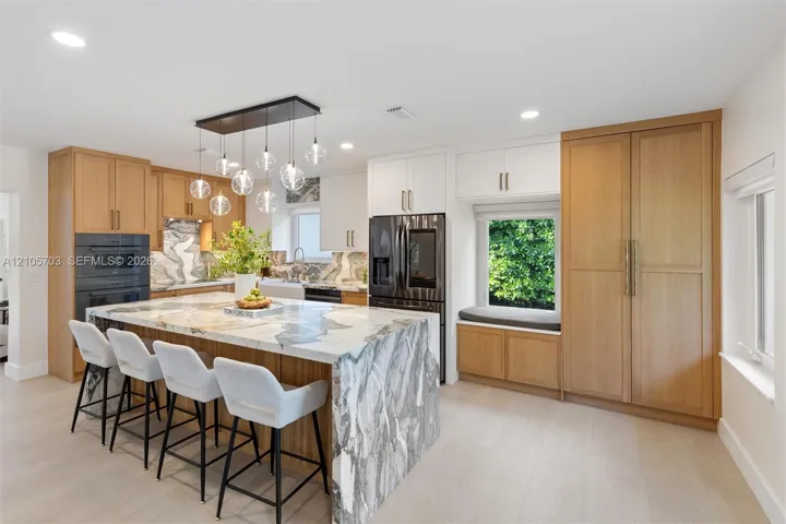 Kitchen featuring a large waterfall island with patterned stone surfacing, natural wood-finish cabinetry, built-in dual ovens, multi-light pendant fixture, and a window-side built-in bench