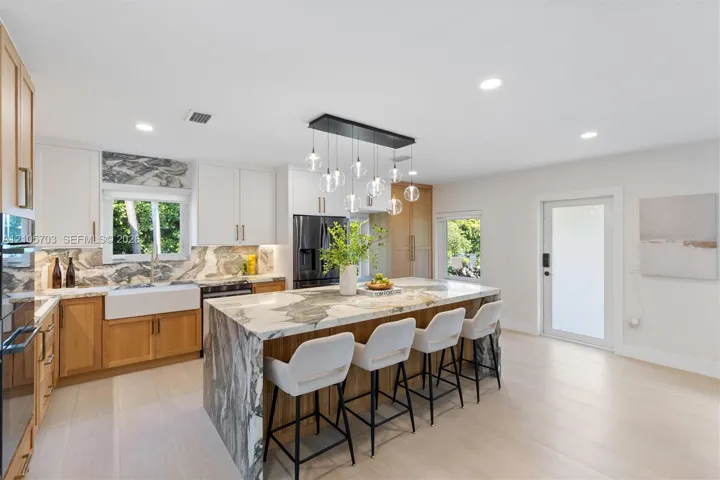 Kitchen featuring a waterfall-edge island with stone surfacing, apron-front farmhouse sink, custom cabinetry with dual-tone finish, built-in stainless steel appliances, and multi-pendant lighting fixture