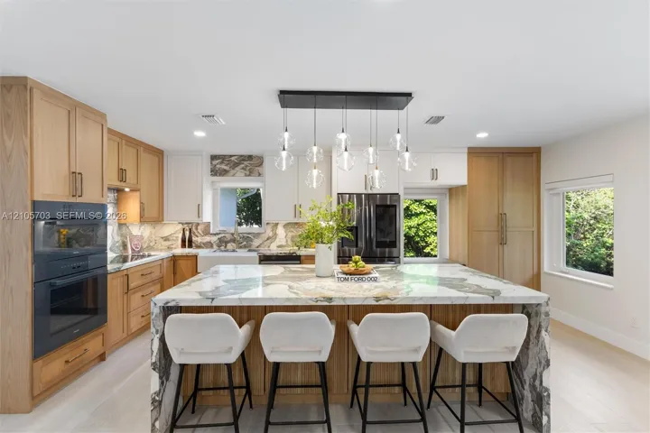 Kitchen featuring custom wood cabinetry, stone-topped waterfall island, built-in double ovens, farmhouse-style sink, and multi-pendant lighting fixture