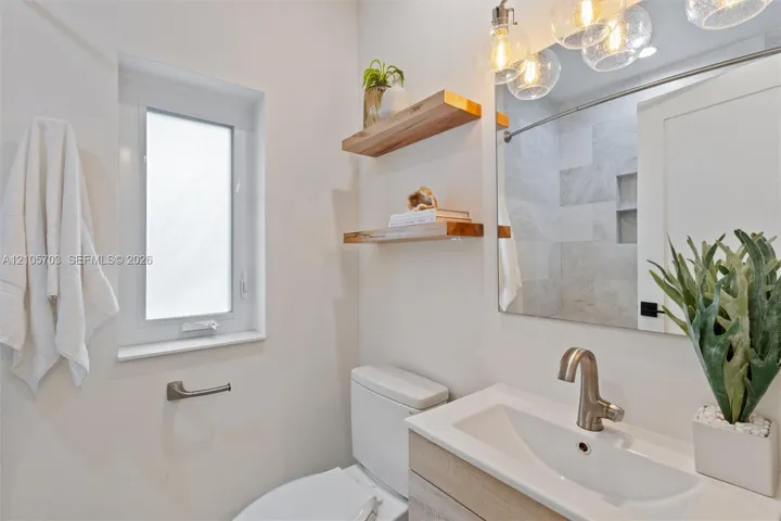 Bathroom featuring a light-toned wood-finish vanity with a rectangular sink, wall-mounted natural wood shelving, multi-bulb globe pendant lighting, and a walk-in shower with tiled surround