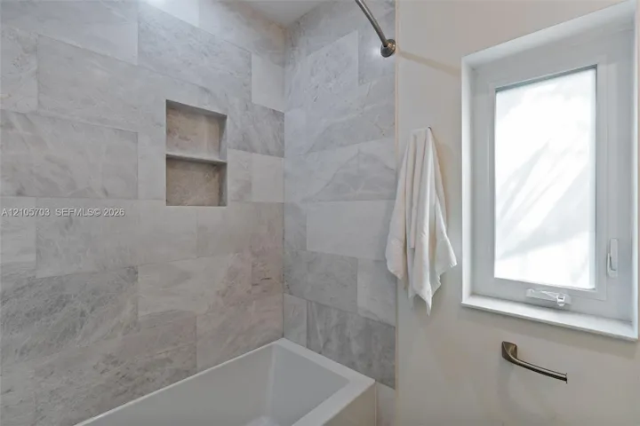 Full-height shower surround featuring large-format light grey stone tiling, recessed double-niche shelving, a built-in soaking tub, and a window with a mounted metal grab bar