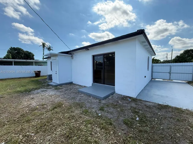 Exterior featuring white stucco siding, sliding glass doors with dark framing, a concrete paved parking area, and a white vinyl perimeter fence