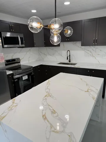 Kitchen featuring dark cabinetry, expansive stone-finish waterfall island and countertops, full-height matching backsplash, stainless steel appliances, and multi-light modern chandelier