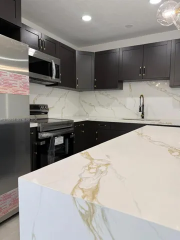 Kitchen featuring dark-toned shaker cabinetry, stone-finish countertops with veining, full-height slab backsplash, stainless steel appliances, and a gooseneck faucet