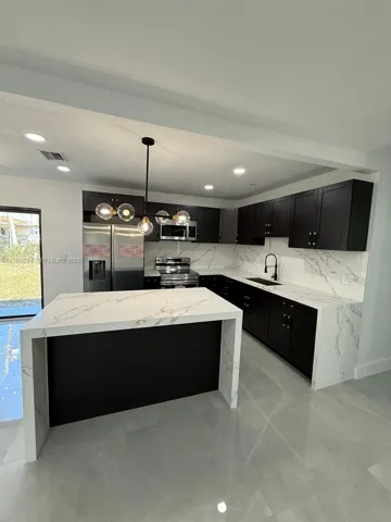 Kitchen featuring dark cabinetry, stone-finish waterfall island, stone-finish backsplash, matte black hardware, and pendant lighting