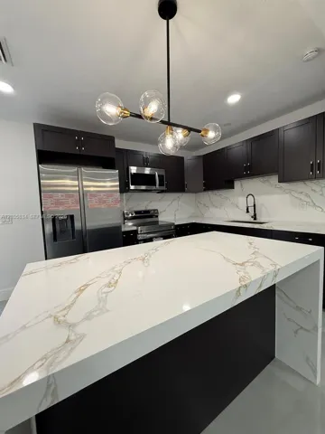 Kitchen featuring an expansive waterfall island with a veined stone-finish countertop, dark cabinetry, stainless steel appliances, and a multi-bulb statement chandelier