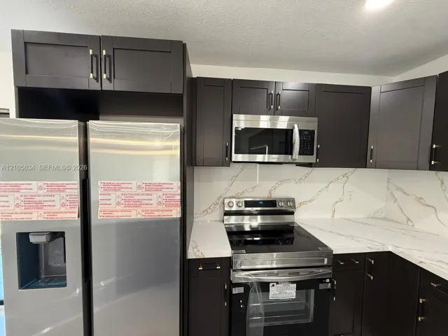 Kitchen featuring dark cabinetry with metallic pulls, stone-patterned countertops, a full-height stone-patterned backsplash, and stainless steel appliances
