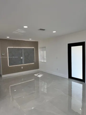 Polished large-format tile flooring, accent wall featuring vertical slat paneling with integrated lighting, centered stone-finish panel, and a black-framed glass door