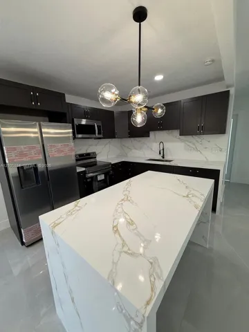 Kitchen featuring dark cabinetry, expansive waterfall-edge island, stone-patterned countertops with matching backsplash, and a contemporary multi-globe pendant light fixture