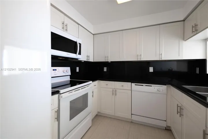 Kitchen featuring white shaker-style cabinetry, black stone countertops, matching black backsplash, and white appliances including a built-in microwave and dishwasher