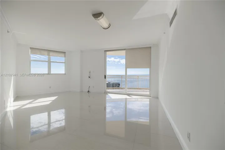 Bright living area featuring large-format polished tile flooring, sliding glass doors to a balcony, neutral wall tones, and expansive windows