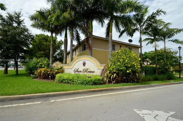 Community entrance featuring a monument sign, manicured lawn, mature palm trees, and native flowering shrubs