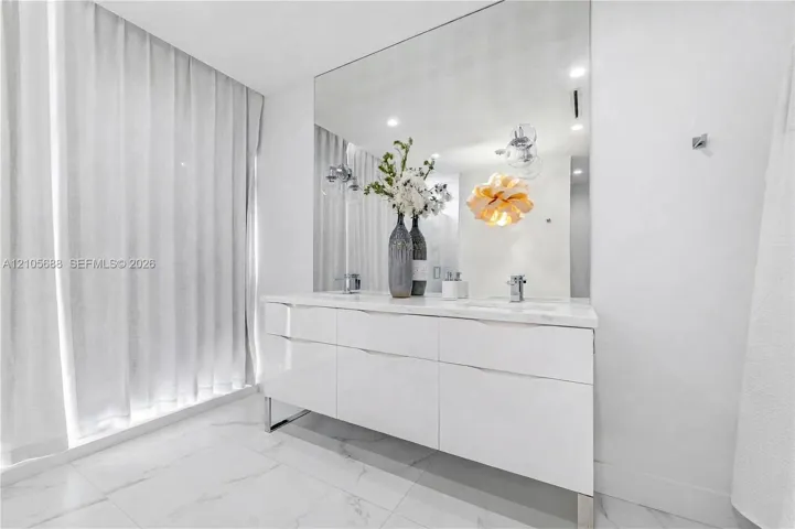 High-gloss white double vanity with an integrated countertop, large-format polished tile flooring, wall-to-wall mirror, and recessed ceiling lighting