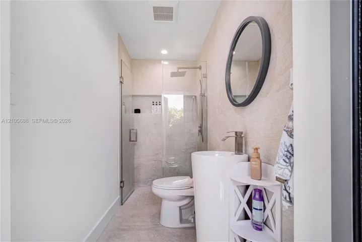 Pedestal sink with metallic faucet, round mirror, glass-enclosed shower, and textured stone-finish wall tile