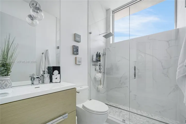 Floating wood-finish vanity with integrated sink, full-height walk-in shower featuring large-format stone-pattern tile, frameless glass enclosure, rainfall showerhead, and upper transom window