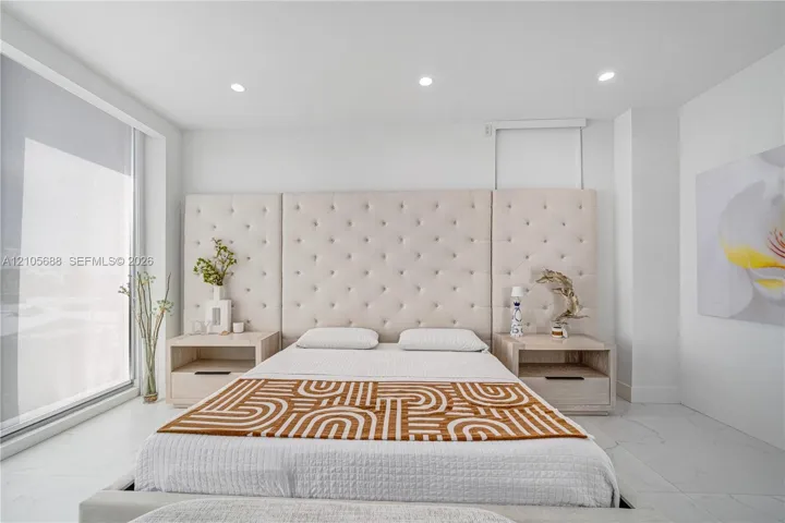 Large-format tile flooring, recessed ceiling lighting, floor-to-ceiling glass window panel, and built-in bedside cabinetry featuring integrated drawer pulls