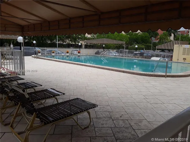 Swimming pool with paved surround, featuring a shaded canopy area and metal perimeter fencing