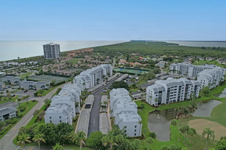 Aerial overview of a multi-building residential complex featuring a water body with a fountain, tennis courts, and covered parking structures