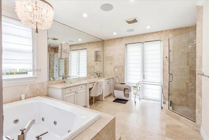 Bathroom featuring a large jetted soaking tub, custom white cabinetry, neutral stone wall and floor tile, glass-enclosed shower, and decorative chandelier lighting