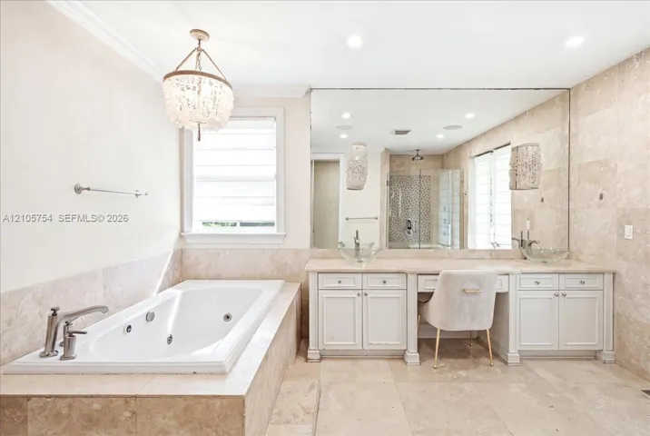 Jetted soaking tub with tile surround, double vanity with built-in dressing area, oversized wall mirror, decorative chandelier, and floor-to-ceiling tile wall finishes
