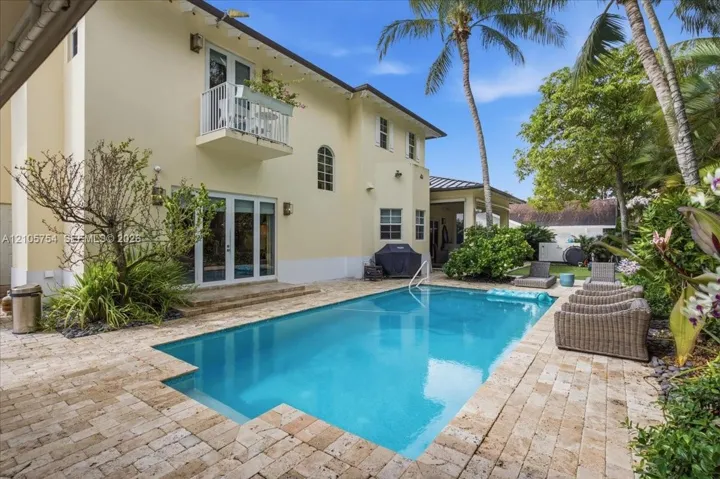 In-ground swimming pool with stone paver decking, second-story balcony with metal railing, double glass-panel doors, and exterior stucco finish