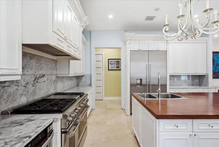 Kitchen featuring white cabinetry, stone countertops with matching backsplash, stainless steel gas range, wood-topped center island with integrated sink, and stone flooring
