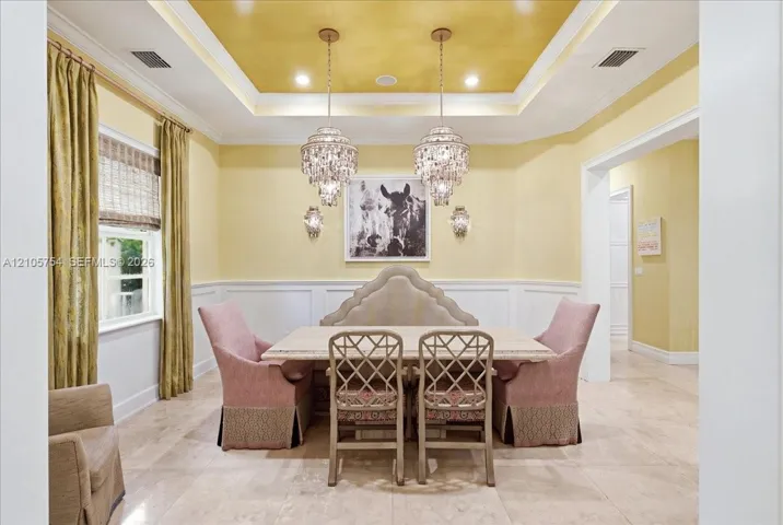 Tray ceiling featuring a decorative metallic finish, dual tiered crystal chandeliers, wall-mounted sconces, wainscoting, and stone-finish flooring