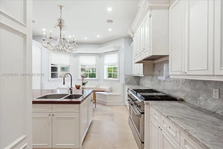 Kitchen featuring custom white cabinetry, stone countertops with matching backsplash, professional-grade stainless steel range, a large island with an integrated double-basin sink, and a built-in window seat
