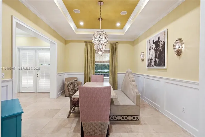 Dining room featuring a double-tiered tray ceiling with gold-tone finish, crystal chandelier, inset wall sconces, wainscoting, and large-format tile flooring