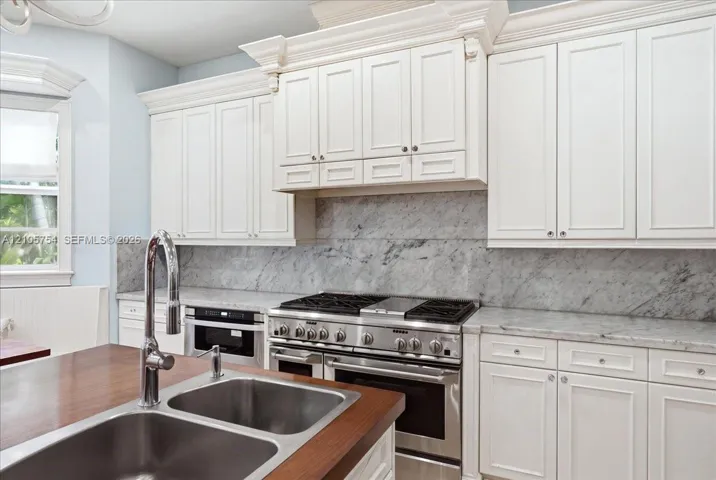 Custom white cabinetry with crown molding, natural stone backsplash and countertops, professional-grade stainless steel gas range, and wood-topped island with an integrated stainless steel dual-basin sink