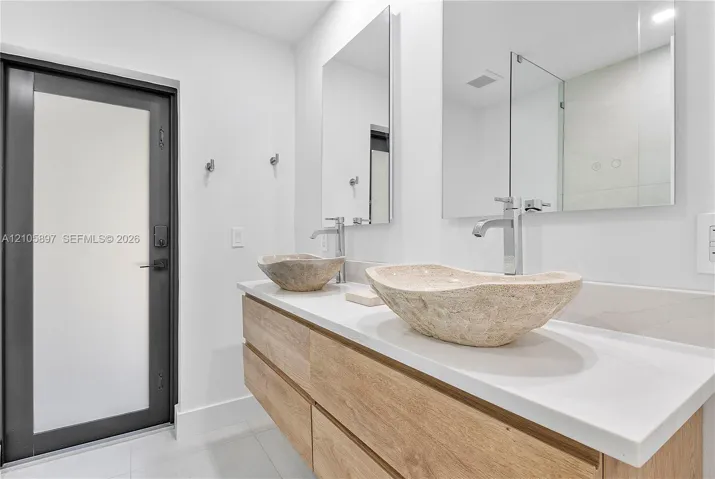 Floating wood-finish vanity with a stone countertop, dual carved-stone vessel sinks, chrome fixtures, and rectangular mirrors