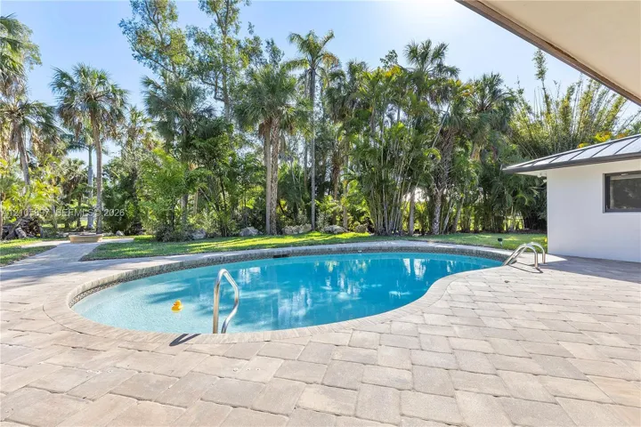 Free-form swimming pool with metal handrails, expansive stone-paver patio, and mature tropical landscaping