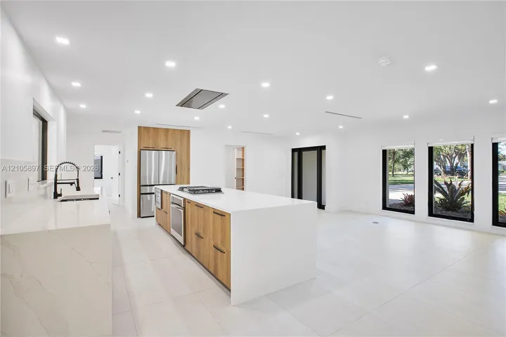 Open-concept kitchen featuring a large waterfall-edge island, wood-finish cabinetry, stone countertops, stainless steel appliances, and recessed lighting