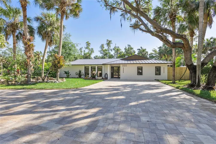 Single-story exterior featuring a metal roof, paver driveway, light-toned stucco facade, and established tropical landscaping