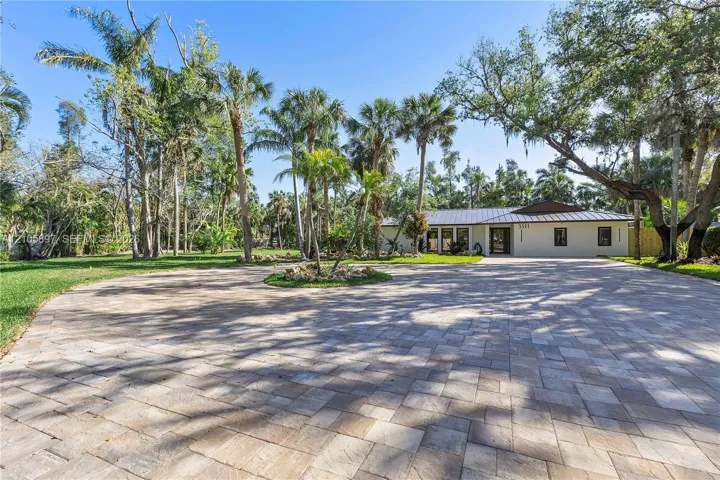 Extensive paver driveway featuring a central circular garden bed, mature palm trees, sprawling lawn, and a light-toned exterior finish with a metal roof