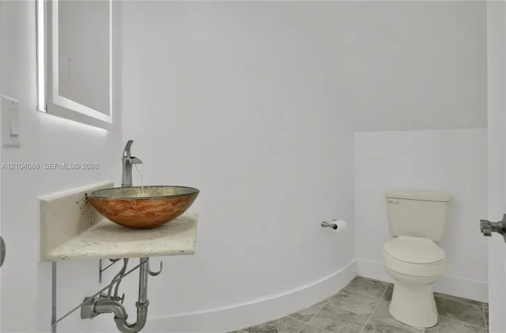 Wall-mounted floating stone vanity featuring a circular copper-tone vessel sink, brushed metal faucet, integrated LED-backlit mirror, and grey tile flooring