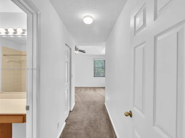 Hallway featuring neutral wall-to-wall carpeting, white six-panel interior doors, and a bathroom vanity with a light-toned countertop and multi-bulb integrated lighting