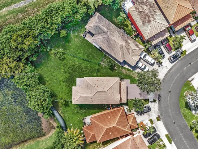 Aerial view of residential property featuring a hipped tile roof, paved driveway, expansive lawn, and mature perimeter trees bordering a natural water feature