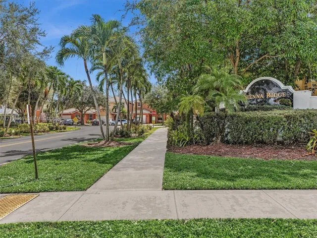 Community entrance featuring a concrete sidewalk, landscaped grounds with manicured lawns, mature palm trees, and established shrubbery