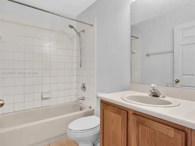 Bathroom featuring a white tile tub surround, handheld shower fixture, wood-finish vanity with an integrated countertop sink, and a full-width vanity mirror