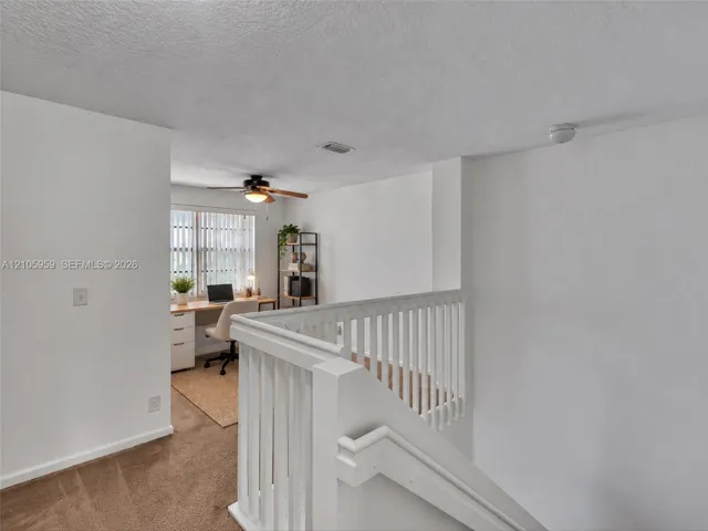 Neutral interior space featuring white walls, carpeted flooring, a white wooden staircase railing, and a ceiling-mounted fan with integrated lighting