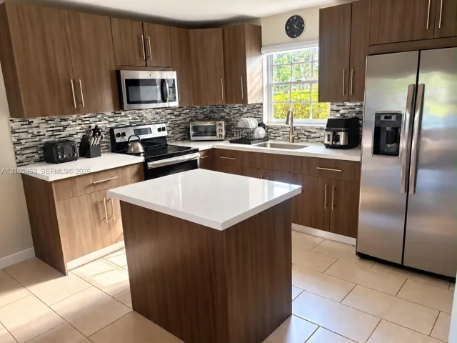 Kitchen featuring wood-finish cabinetry, stainless steel appliances, a center island with a white countertop, a mosaic tile backsplash, and light-toned tile flooring