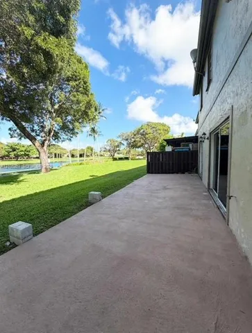 Expansive concrete patio with sliding glass doors, a manicured lawn, mature trees, and a water feature