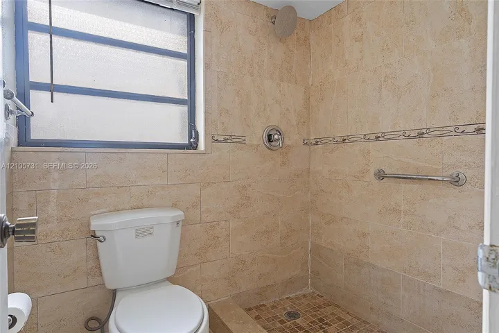 Full-height stone-tile wall finishes, walk-in shower with stone floor tiling, wall-mounted metal grab bar, overhead shower fixture with integrated temperature control, and opaque-glass window
