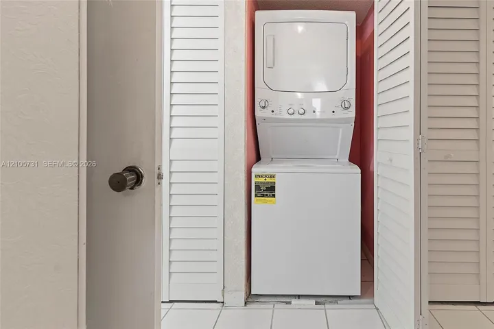 Stacked laundry unit housed within a closet featuring white louvered doors and tile flooring
